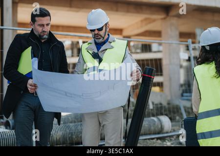 Construction engineers reviewing blueprints at an active building site with hard hats and safety vests Stock Photo