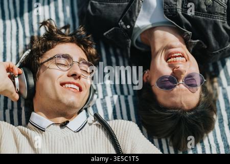 Two friends relaxing outdoors on a striped blanket listening to music with headphones and sunglasses Stock Photo