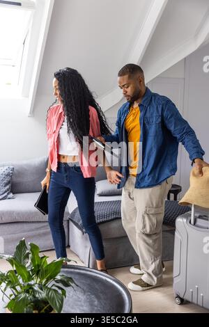 Diverse couple packing grey rolling suitcase, holding straw hat and tablets in loft living room Stock Photo
