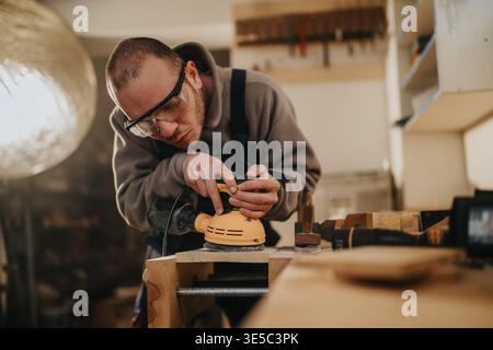 Carpenter sanding wooden plank in workshop focused craftsman using electric sander with safety goggles Stock Photo