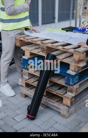 Construction engineers reviewing blueprints on wooden pallets at an outdoor site with plan tube Stock Photo