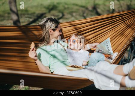 Mother and daughter relaxing in a hammock outdoors sharing snacks and reading a magazine in the park Stock Photo