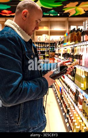 Individual assesses wine in store aisle. Customer carefully examines wine bottle among stocked shelves. Customer in denim jacket thoughtfully inspects Stock Photo