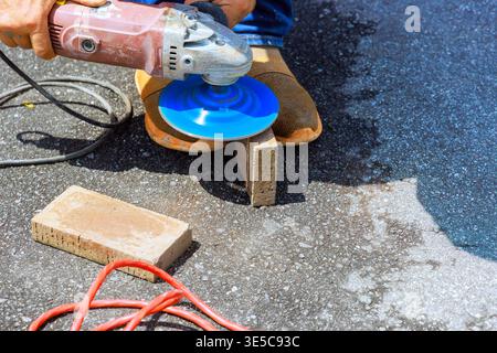 Worker cuts block with grinder tool while standing on dark pavement with other tools, materials around. Stock Photo