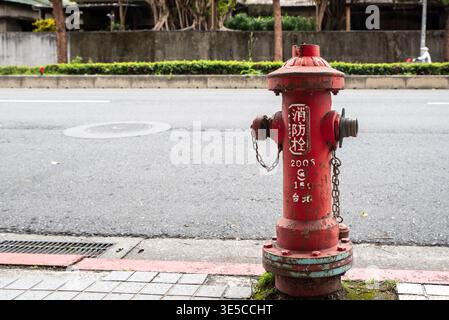 Red fire hydrant stands on paved sidewalk near street. Hydrant shows signs of weathering with moss growth. Chain links hang from caps for safety. Urba Stock Photo