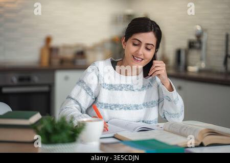Young woman studying at home while talking on phone Stock Photo