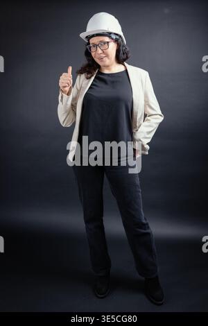 Confident woman engineer wearing hard hat smiling, showing thumbs up on dark background Stock Photo