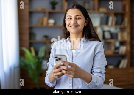 Young indian woman smiling holding a mobile phone in her hands, checking social media, messaging, or browsing online in a comfortable home or office setting Stock Photo