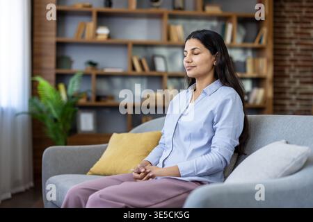 Young woman finding inner peace and balance through meditation exercises, practicing self-care and relaxation on a comfortable couch in her living room Stock Photo