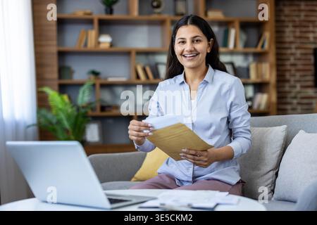 Young smiling woman sitting on sofa at home unpacking a document from a craft envelope with laptop on coffee table, receiving important information or positive results Stock Photo