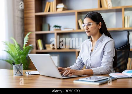 Young indian businesswoman in glasses sits at a modern office desk, focused on typing on her laptop while working remotely in a smart-casual corporate environment Stock Photo