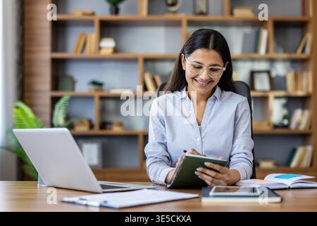 Young professional woman in glasses smiling as she writes in a notebook at a wooden desk with laptop, focused in a modern home office with blurred bookshelf background Stock Photo