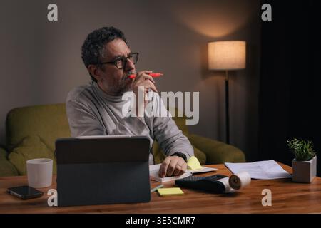 Middle aged man working from home reviewing financial documents and highlighting important information with marker while using tablet. Concept of pers Stock Photo