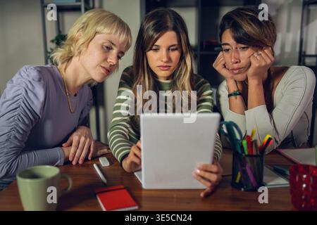 Three young women sitting together at a table while using a digital tablet, smiling and collaborating in an indoor study or creative workspace, modern Stock Photo