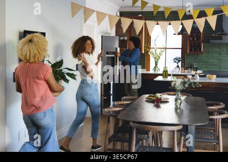 Three adult African American women decorating kitchen with bunting, holding potted plant by fridge Stock Photo
