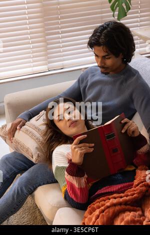 Diverse couple reclining on light-colored sofa near window, woman holding red hardcover book Stock Photo