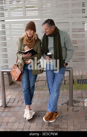 Middle aged couple standing at stop in jackets, woman taking notes in notebook; man holding coffee Stock Photo