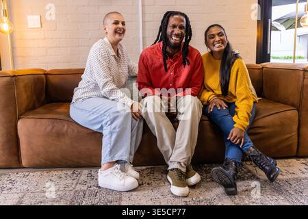 Diverse friends sitting on brown leather sofa in lounge, wearing jeans and lamp glowing Stock Photo