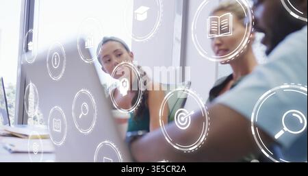 Working three teammates wearing sleeveless and dark tops in office, using laptop with overlay icons Stock Photo