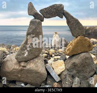 Balanced arrangement of stones on a rocky seashore with calm water and a cloudy sky, showcasing different shapes and sizes of rocks Stock Photo
