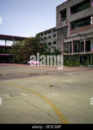 Wuhan, China - June 17, 2025: Abandoned multi-story concrete building surrounded by overgrown weeds and an empty paved parking lot at dusk. The image Stock Photo