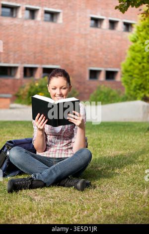 Female student reading book on campus lawn, opening pages and activating animated education notes. Volume, backpack, outdoors, brick, greenery, concen Stock Photo