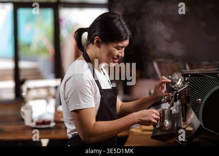 Asian barista in twenties operating espresso machine at coffee shop, frothing milk with pitcher. Coffee, barista, espresso, beverage, café, ambiance, Stock Photo
