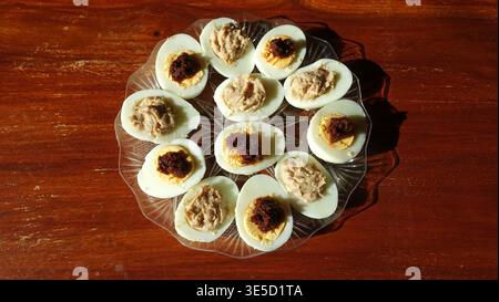 Glass plate of homemade deviled and tuna-stuffed hard-boiled eggs arranged on a rustic wooden table, ideal for brunches, parties, picnics and casual f Stock Photo