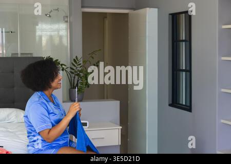Blue satin garment is resting on white bedding near bedside table with clock and plant. Woman, loungewear, silky, bedroom, interior, relaxed, modern Stock Photo