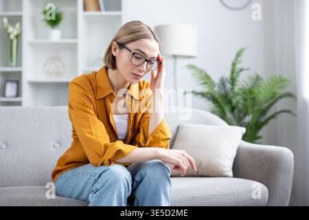 Young woman sitting on a sofa and suffering from a strong headache or migraine, feeling stressed, tired, and experiencing pain while holding her temple Stock Photo
