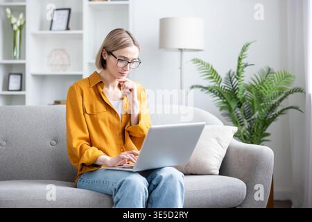 Young woman wearing glasses sitting on a couch, focused on her laptop while telecommuting from her living room, enjoying comfort and flexibility of working from home Stock Photo