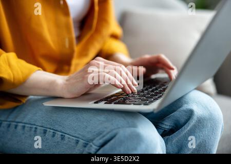 Person wearing a yellow shirt and jeans using a modern laptop for online communication, distant learning, or remote work while sitting casually on a sofa at home Stock Photo