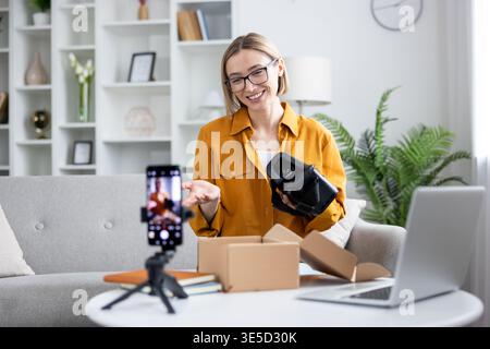 Woman tech vlogger unboxing and demonstrating a new wireless vr headset at home, smiling and recording a live review for her social media channel on sofa, laptop nearby Stock Photo
