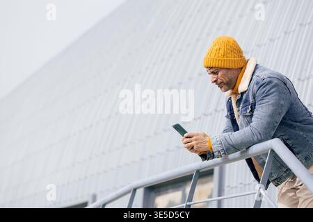 Casual man with a face tattoo leans on a railing, checking his phone while wearing a denim jacket and yellow knit hat in a modern urban setting. Stock Photo