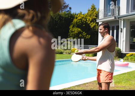 Couple playing paddle-ball with wooden paddles and ball next to backyard swimming pool, copy space Stock Photo