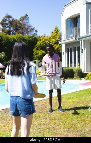 Diverse friends playing paddle ball on backyard lawn, with paddles, green ball and pink pool ring Stock Photo