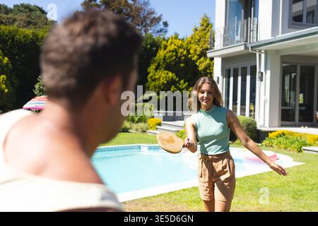 Couple playing paddle game in sunny suburban backyard by pool, with wooden paddles and pool float Stock Photo