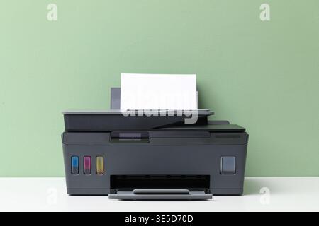 One black printer with sheet of paper on white table near green wall Stock Photo
