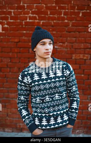 stylish handsome young man with a knitted fashion hat in a blue knitted sweater with a Christmas pattern stands near a red brick wall Stock Photo