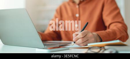 Person Works on Laptop While Taking Notes at a Desk in a Bright Room During the Day Stock Photo