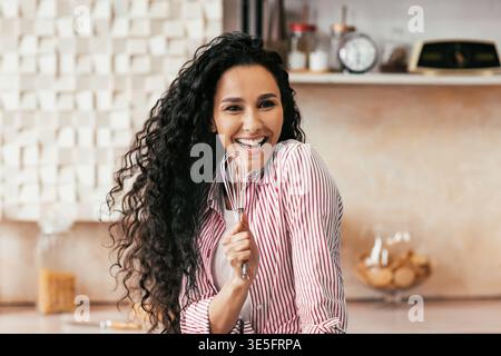 Chef Smiles While Holding a Whisk in a Kitchen With Various Cooking Tools and Ingredients Stock Photo