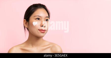 Woman Applying Skincare Product on Face in Front of Light Pink Background While Looking to Side Stock Photo