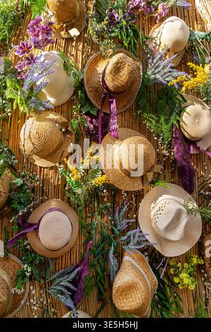 Straw Hats and Flowers on Bamboo Wall. Decorative display of woven straw hats, ribbons, and colorful flowers arranged on a bamboo wall for a cheerful Stock Photo