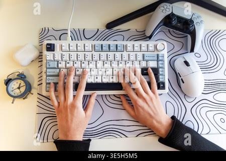 Hands Typing on Modern Gaming Desk Setup. Top view of hands typing on a compact keyboard beside a mouse, game controller, clock, and earbuds case on a Stock Photo
