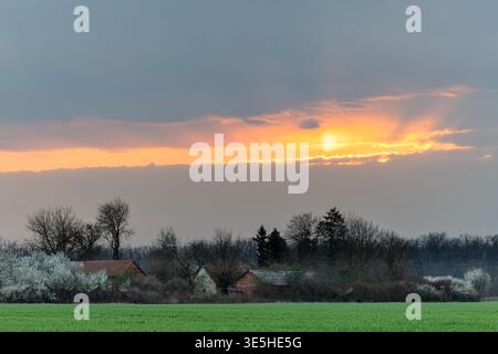 Golden sunset breaks through a dramatic cloud bank above a small rural village, casting warm light across green fields and blossoming trees and creati Stock Photo