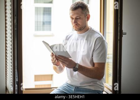 Man reading book while sitting on window sill with natural daylight in modern apartment Stock Photo