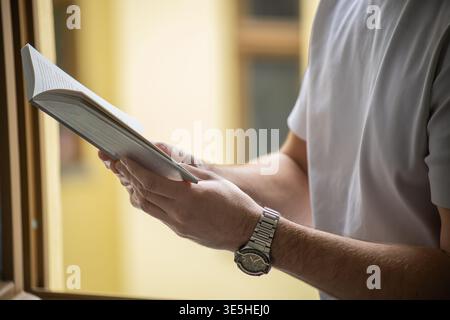Side view of man reading book by window with warm light and shallow depth of field Stock Photo