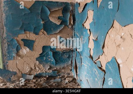 Close-up view of deteriorating blue paint flaking off textured beige wall surface revealing layers beneath in dilapidated structure interior scene Stock Photo