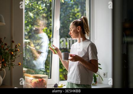 Smiling happy woman reading messages on smartphone while having morning coffee with no rush. Stock Photo