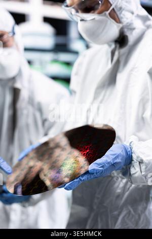 Close up of silicon wafer analyzed by men working together in automated fabrication plant cleanroom. Coworkers in bunny suit examining microelectronic semiconductor substrate for defects Stock Photo